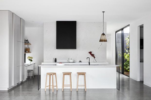 Bright contemporary kitchen with a large white island and integrated sink, three timber bar stools, tall white cabinetry, a white textured feature wall, black rangehood, pendant light, and polished grey concrete flooring. Full-height glazing opens to a leafy outdoor area.