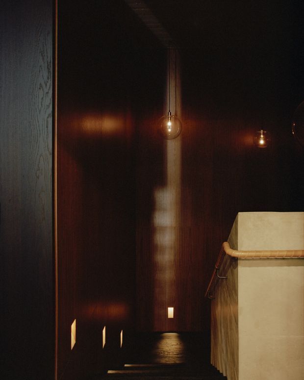 A dark, atmospheric interior passage or stair landing with glossy dark timber wall panelling, a narrow reflective dark floor, warm spherical pendant lights, and small recessed wall lights. A pale sculptural balustrade or partition occupies the right side, while a lighter timber-framed wall edge is visible on the left. The scene has a moody, refined, minimalist architectural character.