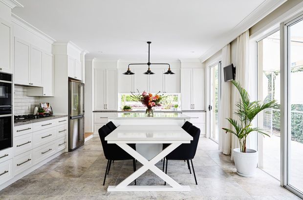 Bright open-plan kitchen and dining space with extensive white shaker-style cabinetry, a central white island, and a white dining table with a bold X-shaped base surrounded by black dining chairs. Large glazed doors and windows bring in abundant daylight, with a potted palm and neutral stone-look flooring adding warmth. Black pendant lights and stainless-steel appliances provide contrast.