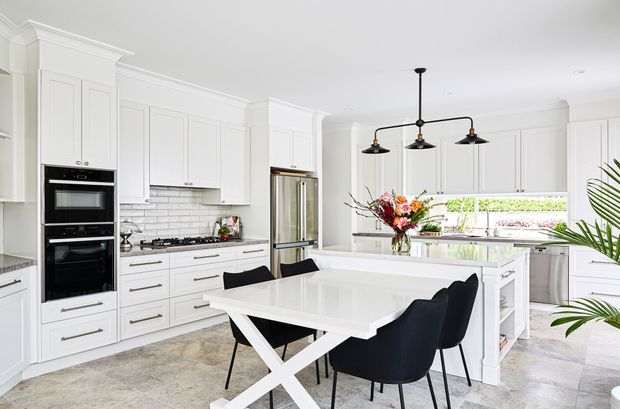 Bright, spacious contemporary kitchen with extensive white Shaker-style cabinetry, integrated black appliances, a large central island with a white benchtop, and a dining table with black upholstered chairs. A linear black pendant light hangs above the dining area, while a tiled backsplash, pale stone-look flooring, stainless-steel refrigerator, and garden-facing window complete the refined interior.