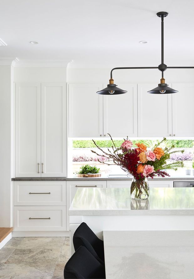 Bright, refined kitchen interior with white Shaker-style cabinetry, a large pale stone island, dark pendant lighting, and a wide horizontal window overlooking landscaped greenery. A floral arrangement sits on the island, with black bar seating partially visible in the foreground.