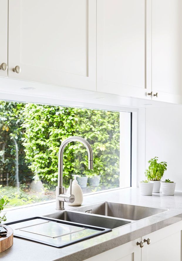 Bright white kitchen sink area with shaker-style overhead and under-bench cabinetry, a large garden-facing window, stainless steel double-bowl sink, curved chrome mixer tap, and pale stone benchtops. Small potted plants sit along the window, creating a fresh, light-filled feel.