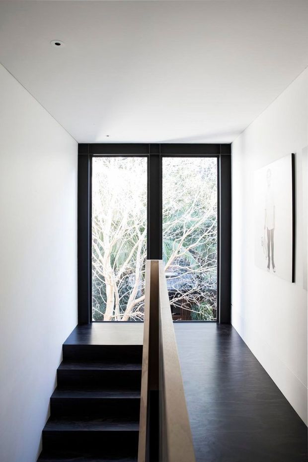 Minimalist interior stairway with dark timber or painted stairs and flooring, white walls and ceiling, and a simple central timber handrail. Full-height black-framed glazing at the landing provides a strong view of trees outside, while a small framed artwork hangs on the right wall. The space is narrow, bright, and carefully detailed with recessed ceiling lights.