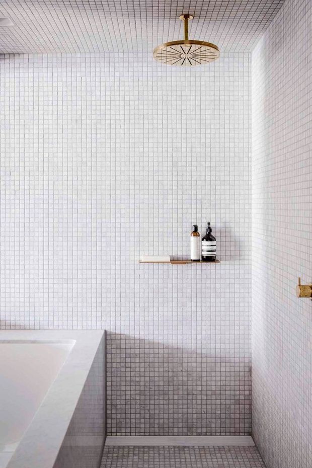 A compact, minimalist bathroom with white square mosaic tiles covering the walls and ceiling, and darker grey mosaic tiles forming the shower floor and lower wall area. A white bathtub is partially visible at left. The walk-in shower has a slim wall-mounted shelf with bottles, a brass shower fixture, and a distinctive brass-and-glass pendant light.