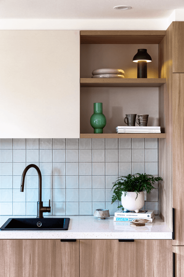 A compact, contemporary kitchen vignette featuring a black undermount sink and curved black tap set into a pale speckled benchtop. Square light blue-grey tiled splashback sits above light timber cabinetry, with open timber shelving displaying ceramics, books, a green glass vase and a small black lamp. A potted fern adds greenery to the benchtop.