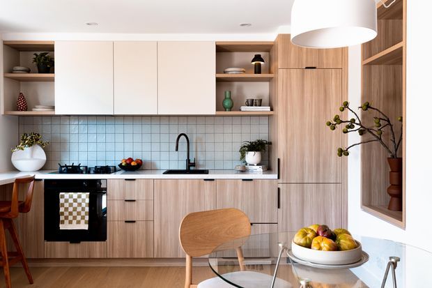 A warm, contemporary open-plan kitchen and dining area with full-height timber-look cabinetry, pale overhead cupboards, open shelving, a tiled splashback, black appliances and tapware, and a compact glass dining table with timber chairs. The space is styled with plants, ceramics, fruit and decorative objects, creating a refined yet inviting atmosphere.