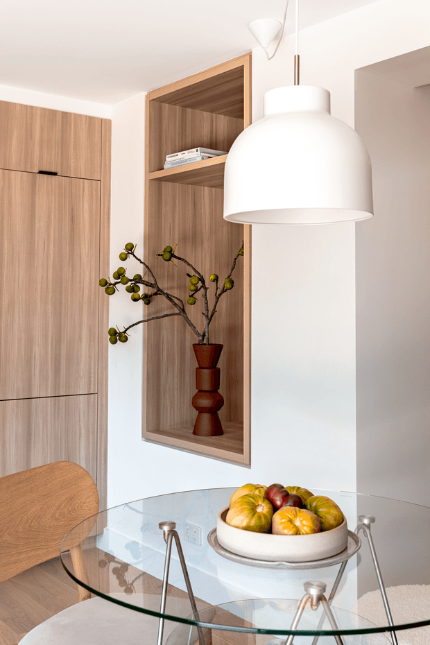 Bright contemporary dining area with a round glass dining table and timber chairs, framed by warm timber built-in cabinetry and an open display niche. A large white pendant light dominates the foreground, while a sculptural vase and bowl of fruit provide decorative accents.