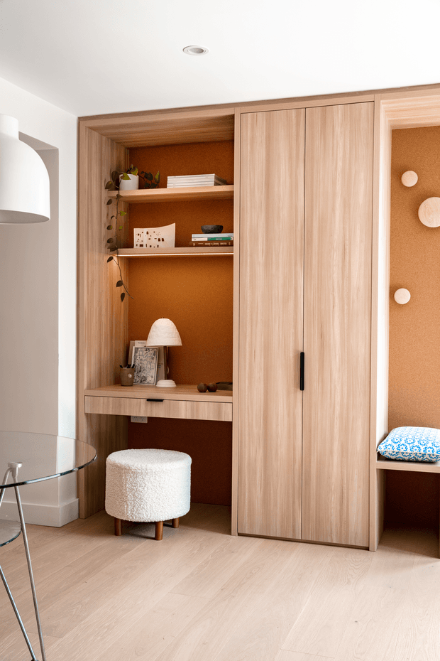 A warm, compact built-in timber study nook and storage wall, featuring open shelving, a built-in desk, tall cupboard doors, and a small upholstered stool. The joinery has a rich timber frame with a textured terracotta-brown backing, while pale timber flooring and white walls create a bright, refined interior. A partial glass table and pendant light are visible at the left.