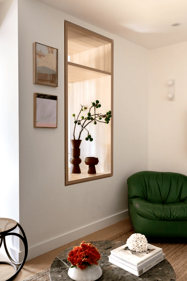 A warmly lit contemporary sitting area with white walls, timber flooring, and a deep green upholstered lounge chair. A large timber-framed interior window or display opening reveals sheer curtains, sculptural ceramics, and a branch arrangement, while two framed artworks hang beside it. A round dark stone coffee table with stacked books and a floral arrangement occupies the foreground.