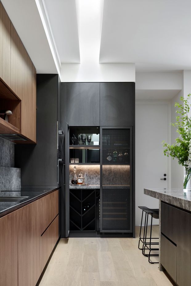 A refined contemporary kitchen with a narrow galley-style layout. The space features dark charcoal cabinetry, warm vertical timber cabinetry and shelving, a dark stone benchtop and splashback, integrated appliances, and a built-in bar or drinks cabinet with textured metal mesh doors and illuminated glassware storage. A central island with bar seating occupies the right side, while pale timber flooring, white walls, and a sculptural linear ceiling detail keep the room bright. A vase of greenery adds a natural touch.