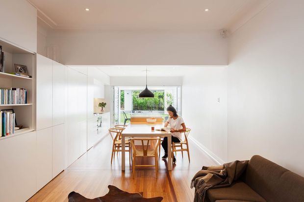 Bright, narrow open-plan dining and living area with extensive white built-in cabinetry and open shelving along one wall, a timber dining table and chairs beneath a black pendant light, and a brown sofa in the foreground. Warm timber flooring, white walls, recessed ceiling lights, and rear glazing create a clean, airy interior.