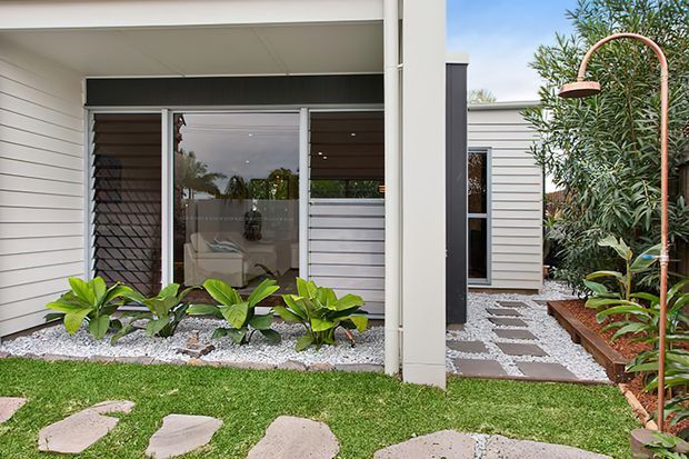Exterior view of a contemporary single-storey home featuring white horizontal weatherboard cladding, dark timber-look window screening, and large glazed openings. A landscaped courtyard includes white gravel garden beds, tropical planting, stepping-stone paths, raised garden edging, and a copper-toned curved outdoor shower or garden feature. A living area is visible through the glazing.