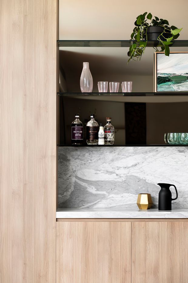 Close-up of a refined built-in home bar and display joinery. Pale timber cabinetry frames dark open shelving, with bottles and glassware displayed above a white-grey veined stone splashback and benchtop. A hanging trailing plant, artwork, ceramic vessels and a black jug add decorative detail.