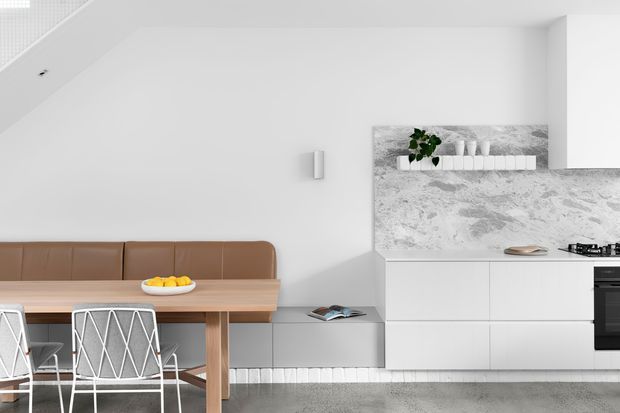 A bright, minimalist open-plan kitchen and dining area with a built-in brown leather banquette, light timber dining table, white geometric wire-frame chairs, and streamlined white cabinetry. The kitchen features a pale grey marble-look splashback and benchtop, integrated appliances, and a small open shelf with a potted plant. A grey carpeted floor and white walls create a clean, restrained palette.