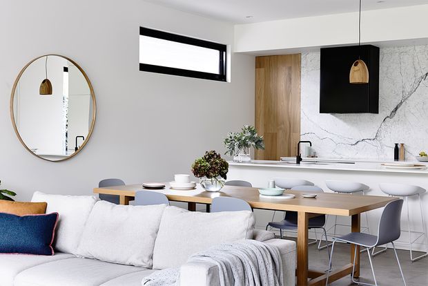 Bright open-plan interior combining a contemporary kitchen, dining area, and living space. The room features white cabinetry and benchtops, a dramatic veined stone splashback, warm timber cabinetry and dining furniture, a black-framed horizontal window, pendant lighting, and a light upholstered sofa in the foreground.