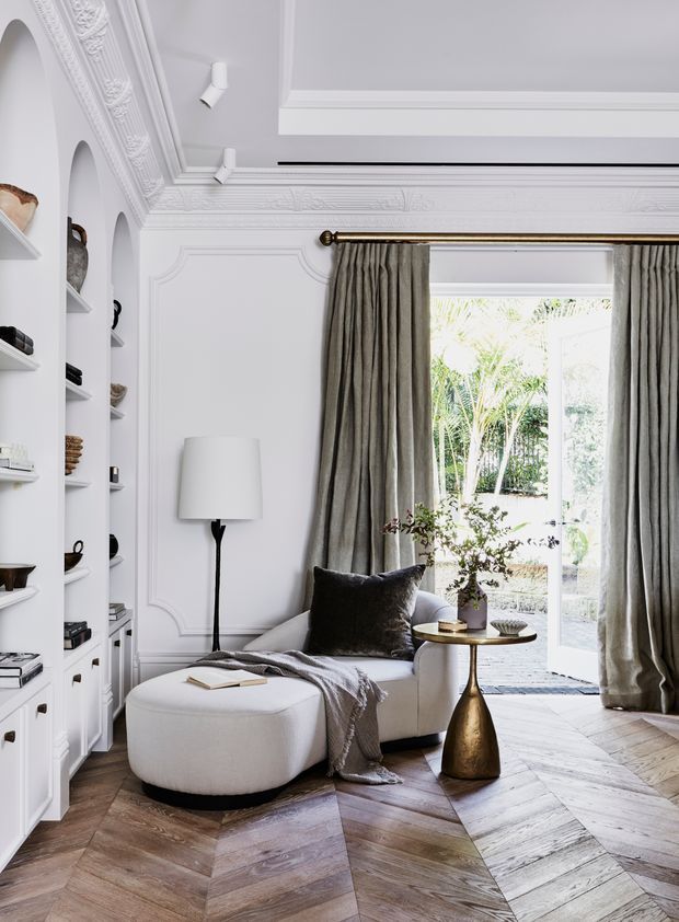 Elegant, refined sitting room with ornate white wall and ceiling mouldings, built-in display shelving, a curved white upholstered sofa and ottoman, layered neutral textiles, and a small sculptural brass side table. Full-height taupe curtains frame glazed garden doors, while the herringbone timber floor adds warmth and pattern.