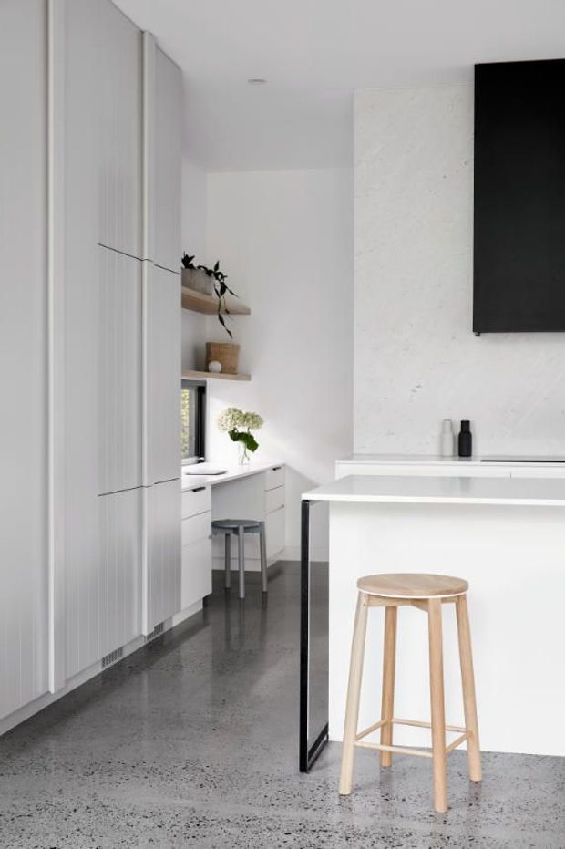 A bright, minimalist kitchen with full-height white cabinetry, a white island with a dark framed side panel, integrated appliances, open timber shelving, and a light timber bar stool. The space features a polished grey concrete or terrazzo-like floor, white walls, and a large black wall-mounted screen or panel.