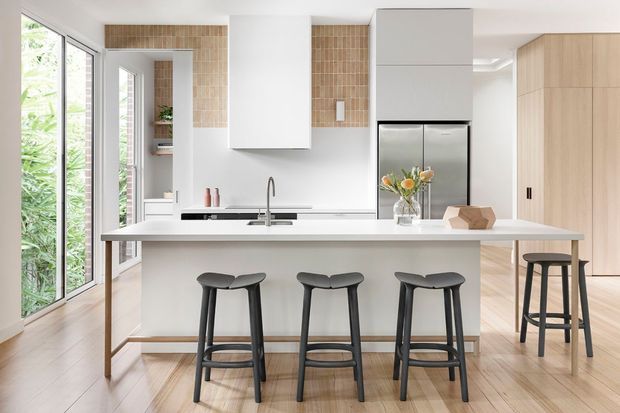 Bright contemporary kitchen with a large white island and integrated sink, three dark stools along the front and an additional bar stool at the side. White flat-panel cabinetry, stainless-steel refrigerator, pale timber flooring and a warm beige tiled splashback are visible, along with full-height glazing opening toward greenery.