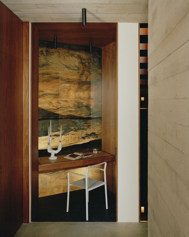 A narrow, richly detailed built-in study nook or writing desk set within dark timber joinery. The alcove features a dramatic natural stone or rock-textured feature wall, a timber desktop with books and a candleholder, a simple white chair, dark carpeting, and slender black pendant lights. A pale textured masonry wall and a black-and-white striped vertical detail frame the space, creating a refined, moody architectural interior.