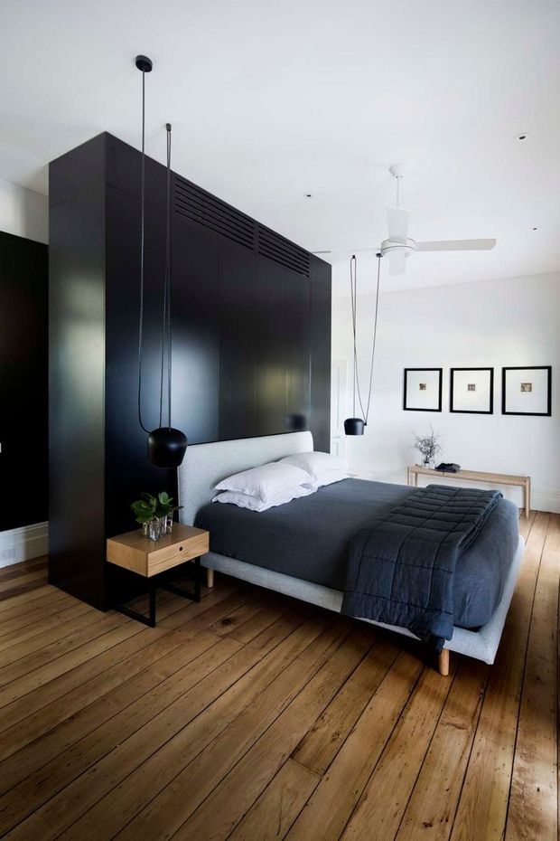 A bright contemporary bedroom with a low upholstered bed dressed in dark charcoal bedding, set against a dramatic floor-to-ceiling matte black built-in wall and partition. Warm timber floorboards, a small timber bedside table, black pendant lights, a white ceiling fan, and three simple framed artworks complete the restrained monochrome interior.