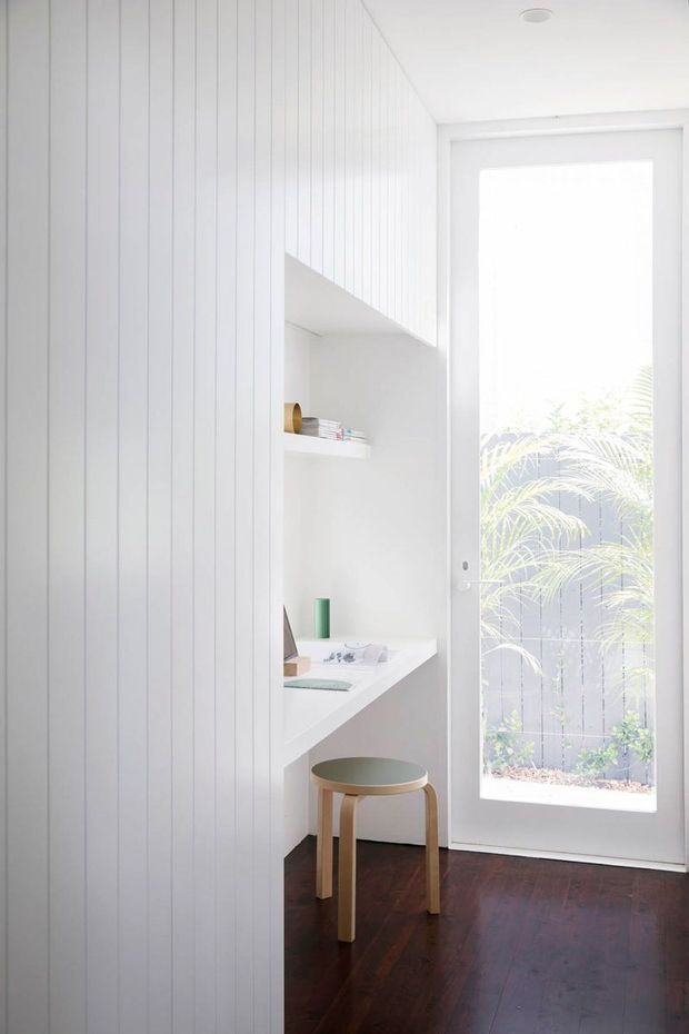 Bright, compact white-lined study nook beside a full-height glazed door. Vertical white wall panelling and built-in white shelving and desk create a minimal workspace, with a simple wooden stool and dark timber flooring. The glazing looks onto a small planted courtyard.