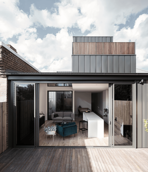 A contemporary rear extension viewed from a timber deck, with large sliding glass doors opening into a bright open-plan living and kitchen area. The home combines dark metal framing, pale brickwork, timber screening and decking, with a raised upper-level volume clad in vertical metal panels and timber battens. Inside are a grey sofa, teal armchair, white kitchen island and pale timber flooring.