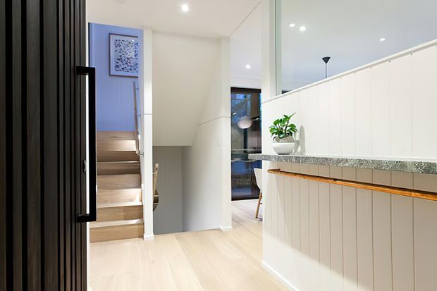 Bright contemporary interior showing an entryway and stair hall with pale timber flooring, a white vertical-panelled kitchen or bar counter with a speckled stone benchtop, black-framed glazing, and a dark vertical-panelled entry door. Timber stairs lead upward beside a white wall and concealed storage.