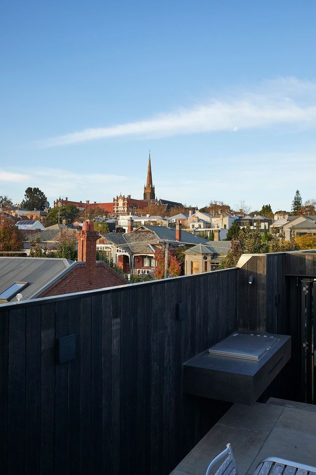 A rooftop terrace or balcony overlooking a low-rise residential neighbourhood, with a prominent historic church spire in the distance. The terrace has dark vertical timber screening or fencing, a built-in dark outdoor barbecue or counter, and grey tiled paving with partial outdoor dining furniture. The scene is photographed in clear daylight with a blue sky and autumnal trees.