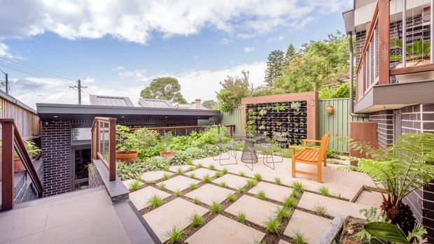 A bright landscaped courtyard terrace between residential buildings, featuring large square concrete pavers with planted joints, raised garden beds, a vertical garden screen, timber privacy elements, outdoor chairs and a small table. Dark brick and timber-clad building facades surround the space, with overhead utility lines and mature greenery visible beyond.