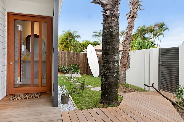 Bright coastal-style residential entry viewed from a timber deck. The image shows a timber-framed glazed front door set within white horizontal weatherboard cladding, opening onto a landscaped courtyard with palm trees, lawn, stepping stones, and a surfboard. A slatted privacy gate and timber decking reinforce the relaxed outdoor setting.