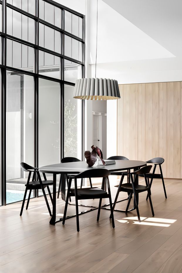 A bright contemporary dining area with a large oval dark dining table surrounded by black dining chairs. A sculptural pendant light hangs above the table, while full-height black-framed glazing creates a strong industrial-style grid along the left side. Pale timber flooring and extensive light timber wall panelling provide warmth, with a glimpse of a blue pool visible beyond the glazing.