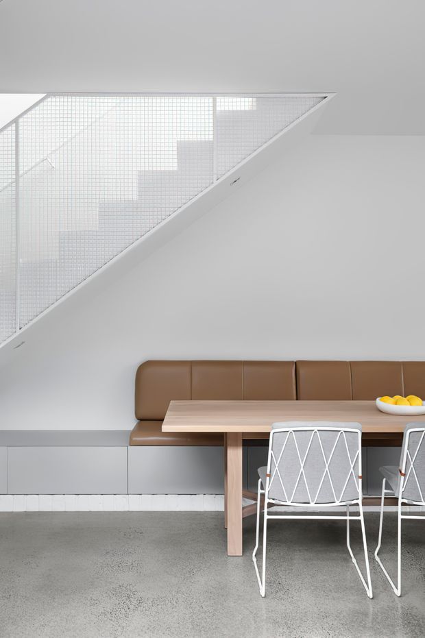 A minimalist dining area with a built-in caramel-brown upholstered bench, pale timber dining table, and white wire-frame chairs. The room features smooth light-grey walls, grey polished concrete flooring, low grey built-in cabinetry, and a striking triangular glazed stair enclosure with a fine square grid pattern. The overall composition is clean, restrained, and modern with soft neutral tones and warm timber accents.