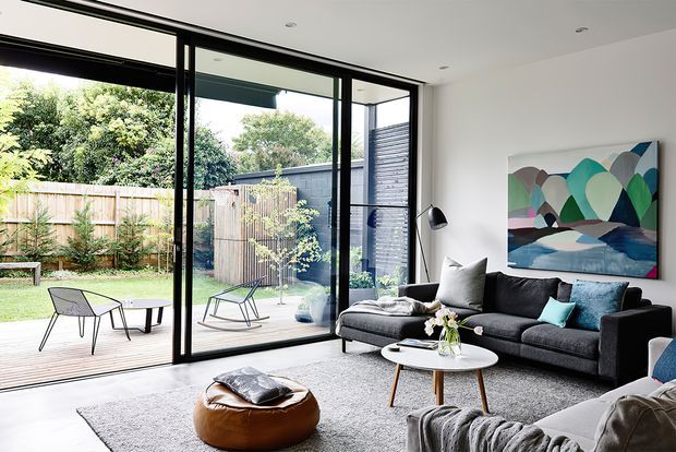 Bright contemporary living room with a dark charcoal sectional sofa, round white coffee table, textured light rug, and colourful abstract artwork. Full-height black-framed sliding glass doors open to a timber deck and landscaped garden, creating a strong indoor–outdoor connection.