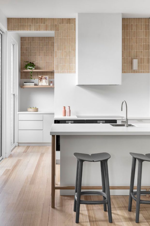 A bright, contemporary kitchen with a white island and integrated cooktop, slim black faucet, pale timber flooring, white cabinetry, and warm beige vertical tiles forming a backsplash and feature wall. Two dark grey bar stools sit at the island, while open shelving and a tall glazed door add depth and natural light.