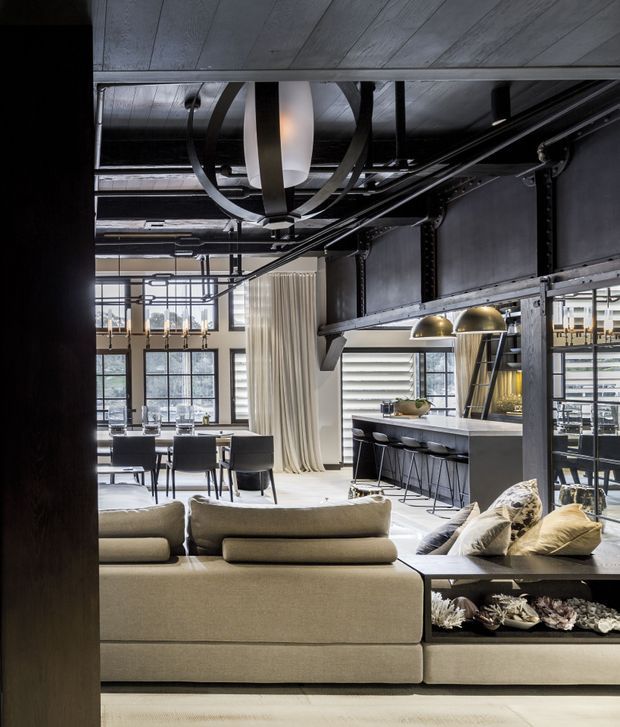 Open-plan loft-style interior combining a living room, dining area and kitchen. The space features exposed dark steel structural framing, exposed services, large industrial-grid windows, a concrete-look ceiling, a neutral sectional sofa, dark dining furniture, a stone kitchen island with bar seating, and sculptural brass pendant lighting. Full-height curtains and layered furnishings soften the otherwise industrial shell.