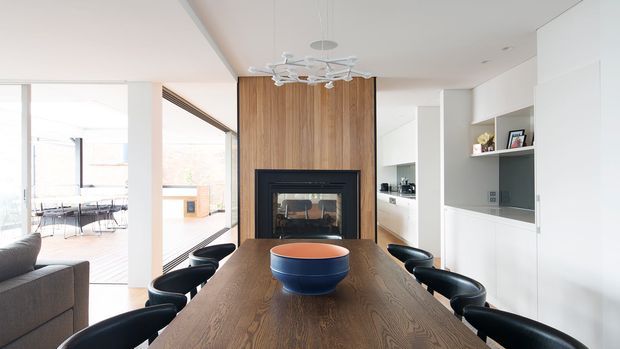 Bright contemporary open-plan dining area with a large dark timber dining table and black curved chairs. A vertical timber feature wall with a built-in fireplace anchors the room, while white cabinetry and a compact kitchen are visible to the right. Full-height glazing opens to a deck or outdoor area on the left, and a sculptural white pendant light hangs above the table.