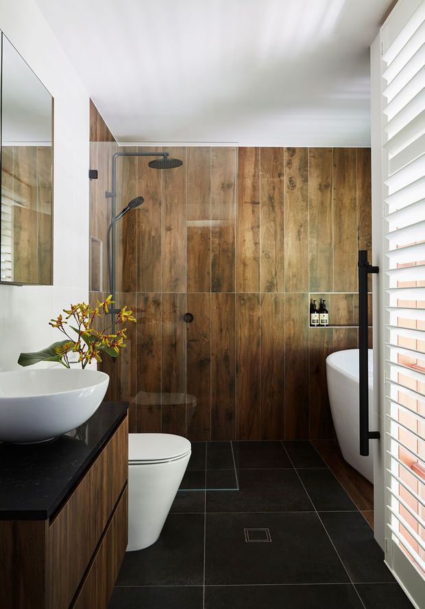 Compact contemporary bathroom with a white vessel basin on a dark benchtop, timber-look vertical wall tiles, dark floor tiles, wall-hung toilet, freestanding bathtub, frameless shower glazing, black tapware and hardware, and white plantation shutters. A mirror and warm timber cabinetry reinforce the refined, natural material palette.