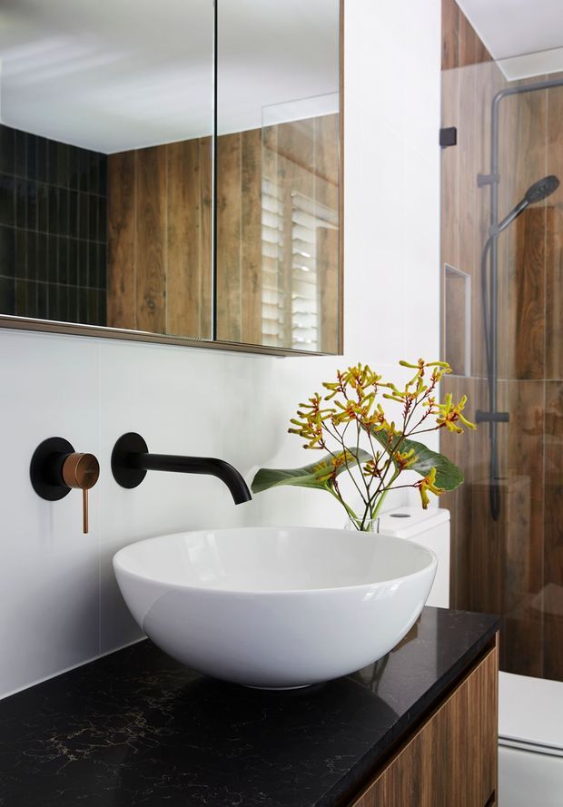 Contemporary bathroom vanity featuring a white vessel basin on a black stone benchtop, wood-grain cabinetry and wall cladding, matte black and bronze tapware, a large mirror, and a framed glass shower enclosure.