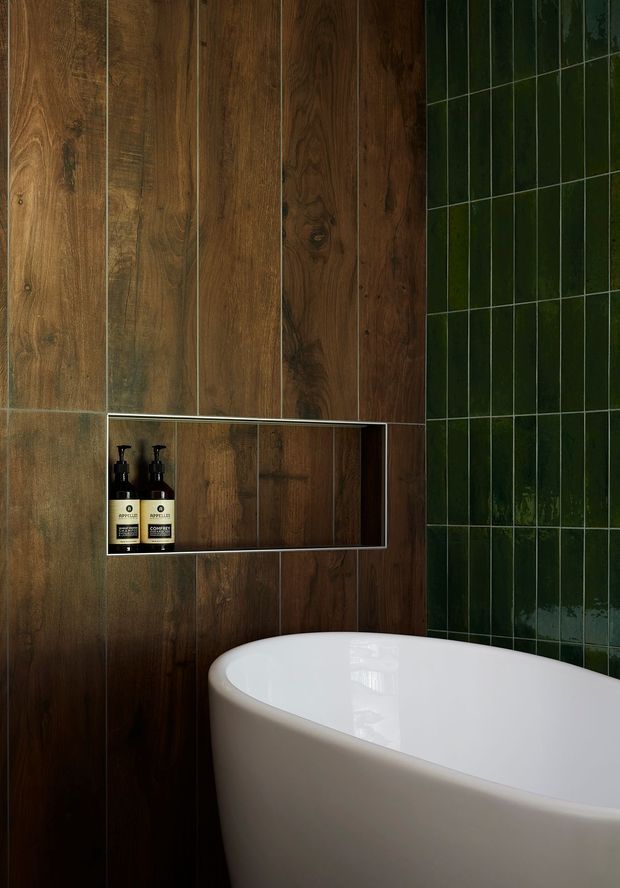 Close-up of a contemporary bathroom featuring a freestanding white bathtub, dark vertical timber wall panelling with an integrated recessed shelf, and glossy dark green rectangular wall tiles.