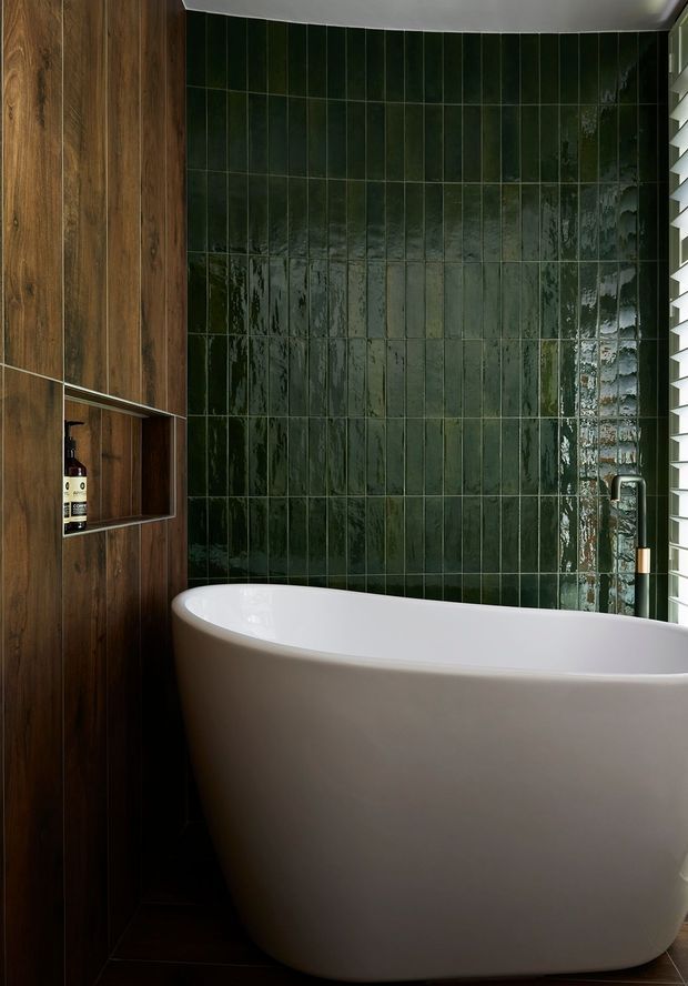 A compact, refined bathroom featuring a freestanding white bathtub against a wall of glossy deep-green vertical tiles. Dark timber wall panelling and built-in open shelving add warmth, while a black and brass tap set and white louvred window provide contrast. The space has a moody, luxurious contemporary feel.