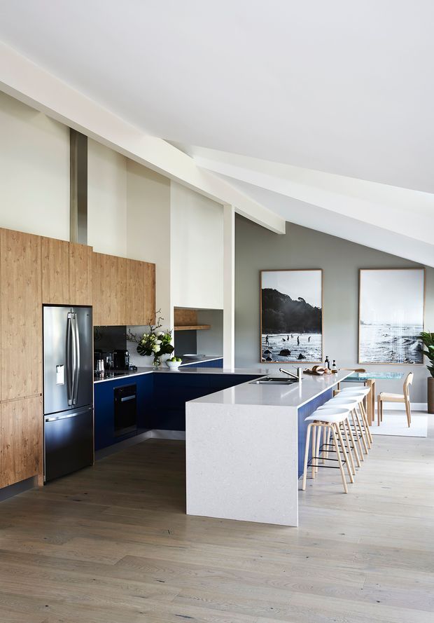 Bright open-plan kitchen and dining area with a large white stone island, deep navy-blue cabinetry, integrated stainless-steel refrigerator, timber wall paneling, pale timber flooring, and white bar stools. The space opens toward a dining table and is decorated with framed coastal artwork and restrained contemporary furnishings. A sculptural angled white ceiling adds architectural interest.