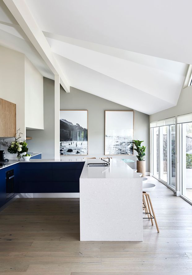 Bright contemporary kitchen with a large white island and integrated seating, deep navy-blue cabinetry, pale grey walls, timber flooring, and expansive glazed doors along one side. The room features a dramatic sloped white ceiling with exposed angular beams, minimalist pendant lighting, framed artwork, and indoor plants.