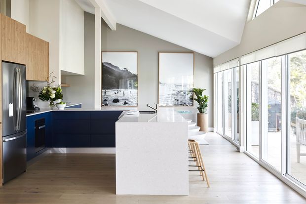 Bright, open-plan contemporary kitchen with a large white island, dark navy-blue lower cabinetry, light timber wall cabinetry, stainless steel appliances, and extensive floor-to-ceiling glazing along one side. The room has a high sloping ceiling, timber flooring, white walls, bar seating, and views to an outdoor terrace.