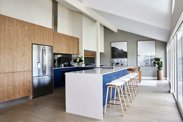 Bright contemporary open-plan kitchen with a large white island, deep blue cabinetry, timber wall panelling, pale timber flooring, stainless-steel refrigerator, and a row of timber-and-white bar stools. Floor-to-ceiling glazing along one side and high angled ceiling planes create an airy, spacious atmosphere.