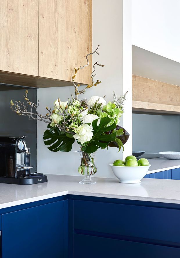 A bright, refined kitchen interior featuring deep blue lower cabinetry, pale timber overhead cabinetry and shelving, a light stone-look benchtop, and a muted blue-grey splashback. A coffee machine, bowl of green apples, and a large floral arrangement sit on the island benchtop. A partial reflection or opening reveals additional blue cabinetry and timber detailing.
