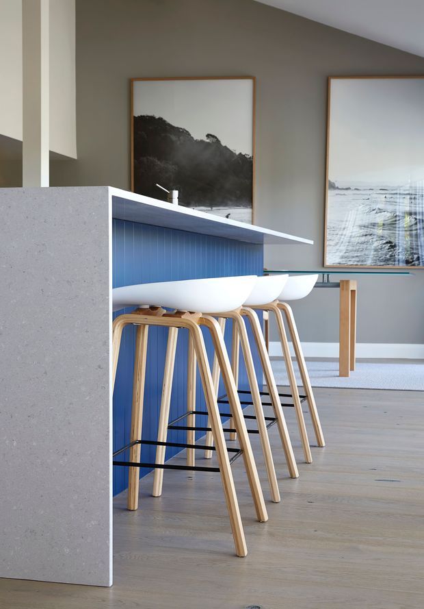 A contemporary open-plan kitchen and dining area with a long blue island featuring a pale stone benchtop and integrated white bar seating. Light timber stools, pale timber flooring, neutral walls, and large framed monochrome coastal and landscape artworks create a clean, bright interior.