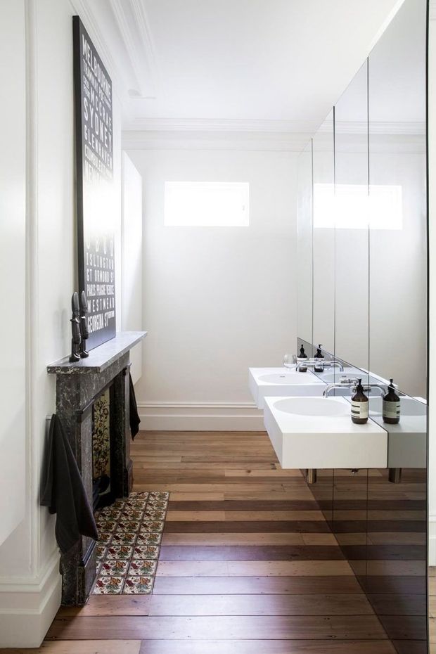 Bright, narrow bathroom with a double floating vanity, wall-to-wall mirrored joinery, polished chrome taps, white walls with ornate cornices and skirting, and warm dark timber floorboards. A dark stone fireplace mantel or console with decorative patterned tiles adds a traditional architectural detail, while the frameless mirror and streamlined basins create a contemporary contrast. Small high windows bring in abundant natural light.