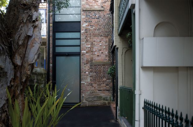 A narrow urban external walkway or laneway between closely spaced buildings. The scene features an aged exposed brick wall, tall dark-framed translucent glazing, painted rendered masonry with an arched recessed detail, a green metal fence, and established planting including a large tree. The architecture has a restrained contemporary intervention alongside traditional masonry fabric.