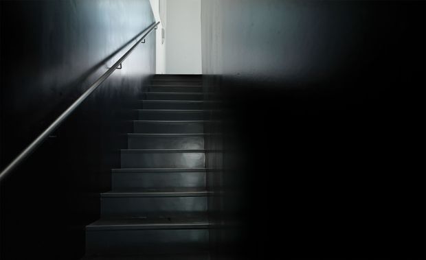 A narrow, dark stairway ascending toward a brighter landing. The stairs and surrounding walls are finished in near-black tones, with a simple metal handrail running along the left wall and minimal detailing. The image has a stark, moody, high-contrast atmosphere.