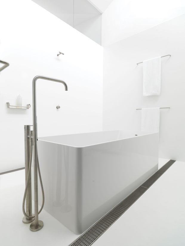 Minimalist white bathroom featuring a large freestanding rectangular bathtub, a sculptural brushed-metal floor-mounted bath filler with hand shower, wall-mounted shower fittings, and simple metal towel rails. The room has seamless white surfaces, a linear floor drain, and bright high-key lighting.